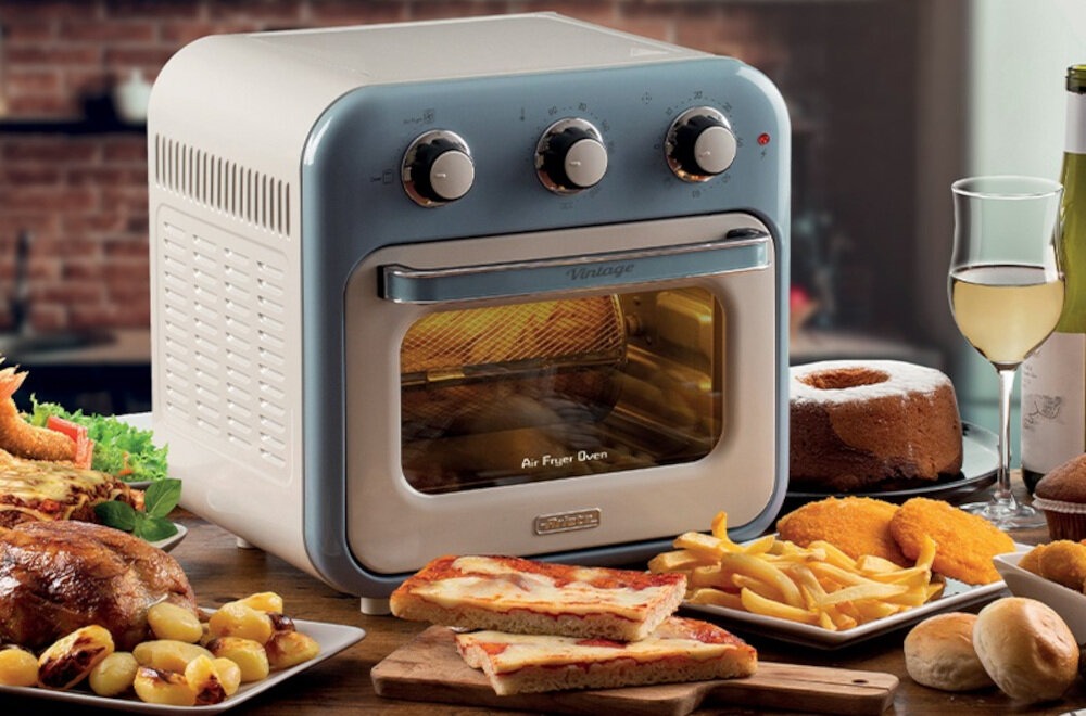 Na obrazie widoczny jest niebieski mini piekarnik z trzema pokrętłami na górze i napisem \'Air Fryer Oven\' na drzwiczkach. Wokół piekarnika znajdują się różne potrawy, takie jak frytki, pizza i ciasto.
