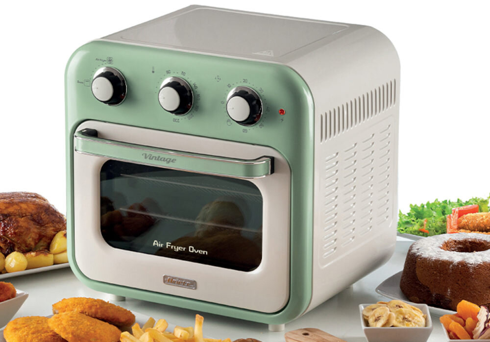 Mini piekarnik w stylu vintage z trzema pokrętłami na górze i napisem \'Air Fryer Oven\' na drzwiczkach. Wokół piekarnika znajdują się różne potrawy, w tym pieczone mięso i ciasto.