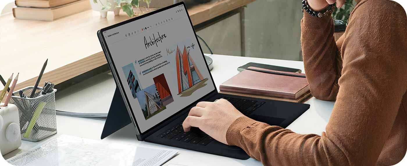 Osoba siedzi przy biurku i używa tabletu z klawiaturą, na ekranie widoczny jest projekt architektoniczny z tekstem \'Architecture\'. Obok leży notatnik i długopis.