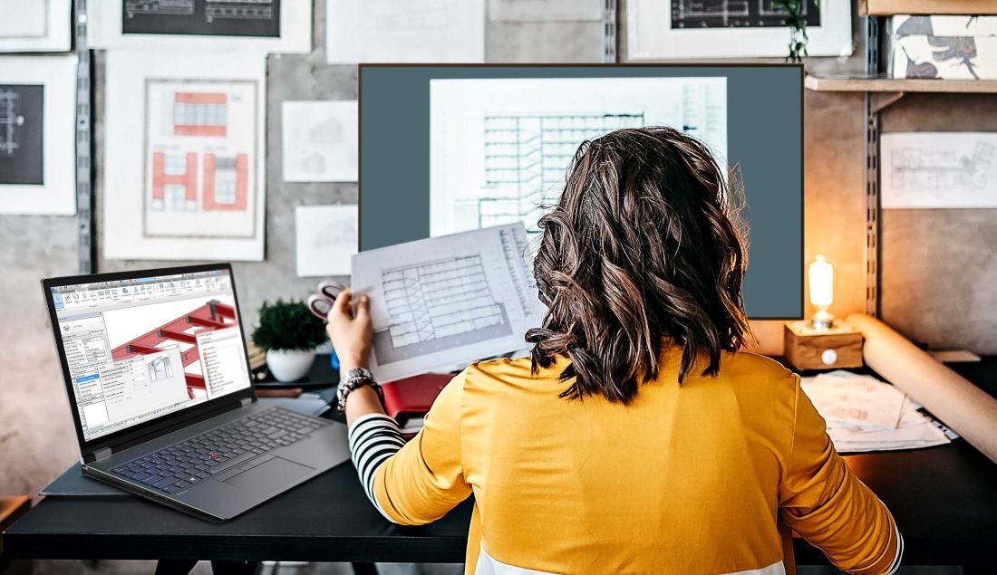 Kobieta w żółtej bluzce pracująca na laptopie Lenovo ThinkPad w biurze architektonicznym, widoczne plany i projekty na biurku - Opis serii ThinkPad P