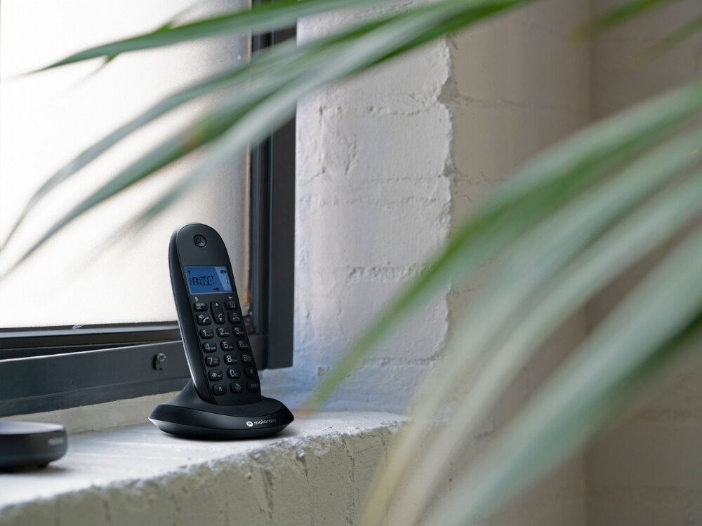Czarny telefon bezprzewodowy stoi na parapecie przy oknie, z wyświetlaczem pokazującym tekst. W tle widoczna jest biała ściana i zielone liście rośliny.