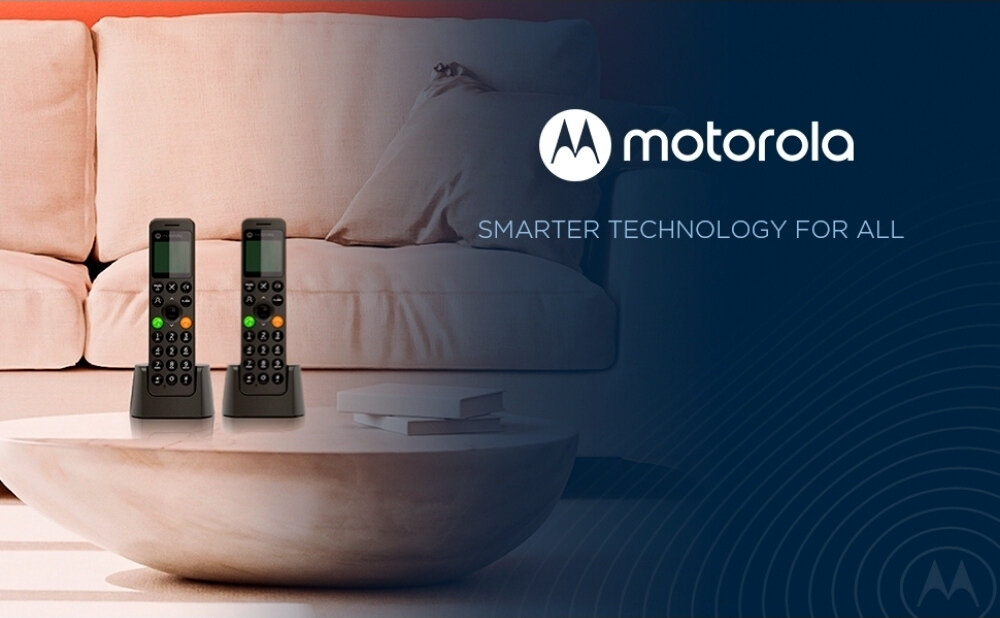 Na obrazie znajdują się dwa czarne telefony bezprzewodowe stojące na okrągłym stoliku przed jasną sofą. Po prawej stronie widoczne jest logo \'motorola\' oraz tekst \'SMARTER TECHNOLOGY FOR ALL\'.