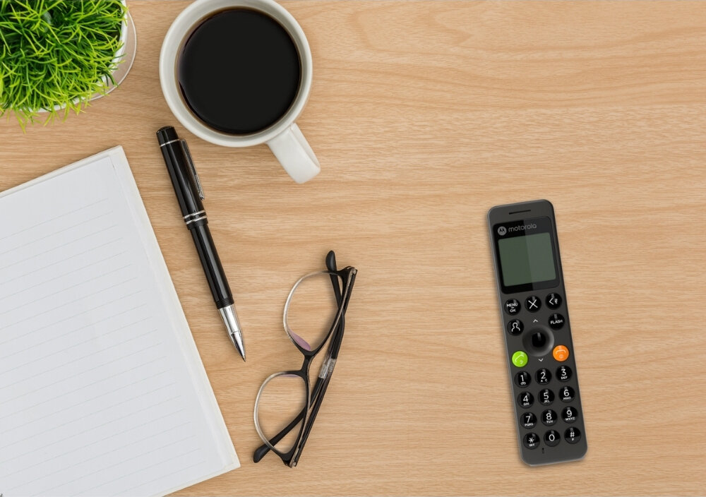 Na drewnianym blacie znajduje się czarny telefon z logo \'motorola\', obok leżą okulary, długopis, notes, filiżanka kawy i mała roślina w doniczce.