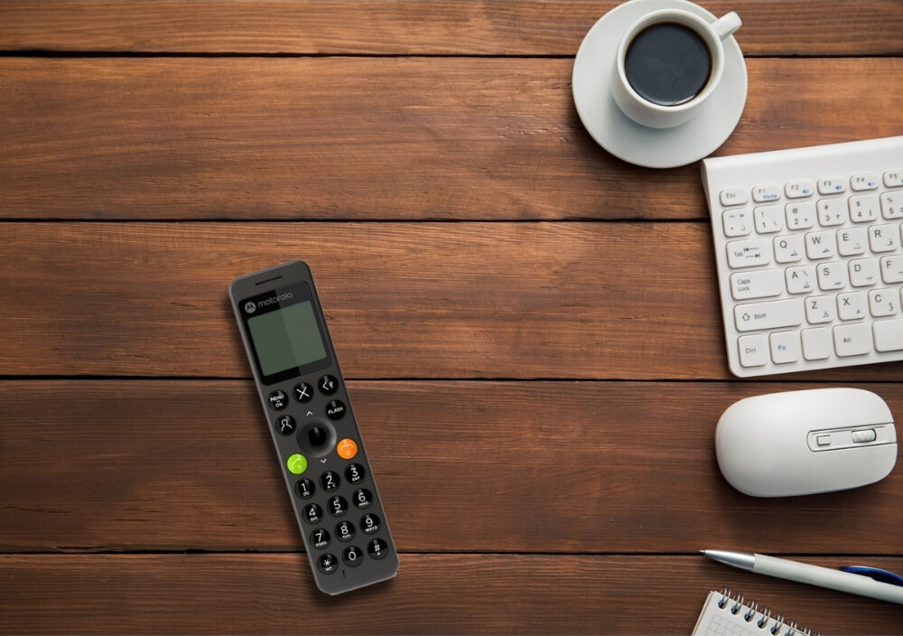 Na drewnianym stole leży czarny telefon bezprzewodowy z widocznym logo \'motorola\', obok znajduje się filiżanka kawy, klawiatura, myszka i długopis na notatniku.