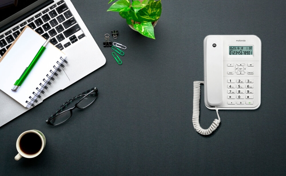 Na biurku znajduje się biały telefon stacjonarny z wyświetlaczem i klawiaturą numeryczną, obok laptopa, notatnika, długopisu, okularów, spinaczy, filiżanki kawy i rośliny doniczkowej. Na telefonie widoczne jest logo \'motorola\'.