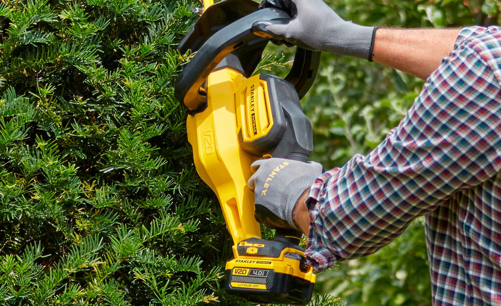 Nożyce do żywopłotu STANLEY SFMCHT855B-XJ 550mm 18V akumulatorowe ogród osoba trzyma nożyce w dłoniach zbliżenie na uchwyt wymagają użycia obu rąk do uruchomienia nożyc minimalizuje ryzyko przypadkowego włączenia ostrza bezpieczeństwo użytkownika
