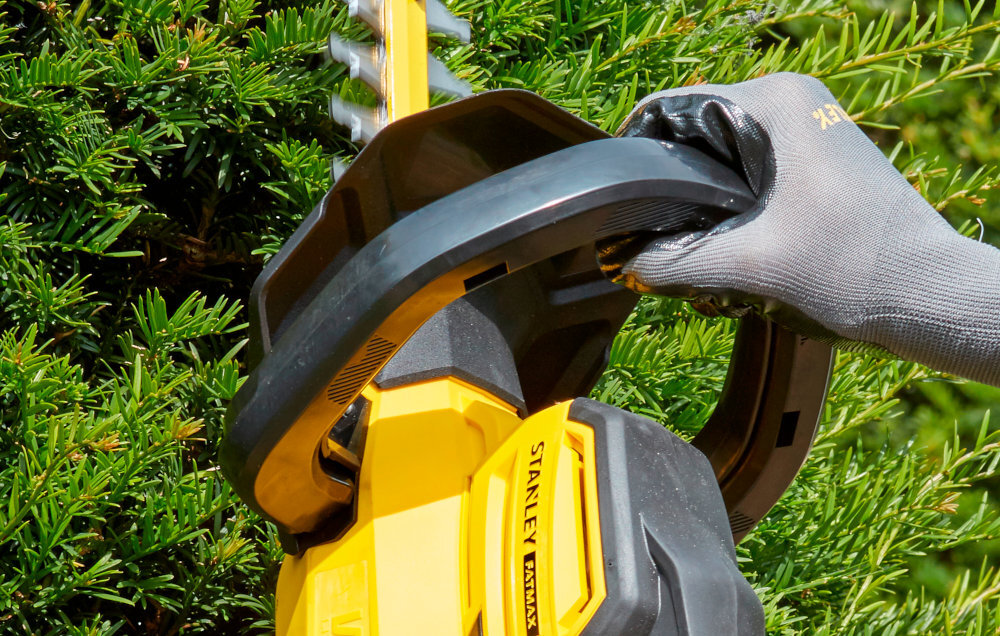 Nożyce do żywopłotu STANLEY SFMCHT855B-XJ 550mm 18V akumulatorowe ściana żywopłotu zbliżenie na osłonę osoba trzyma nożyce w dłoniach osłona chroni dłonie przed odpadkami i gałęziami zwiększa bezpieczeństwo swobodnie manewrować minimalizując ryzyko skaleczeń
