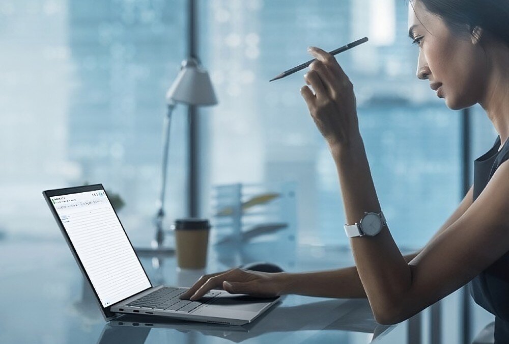 Laptop ASUS ExpertBook CX5403CMA-QM0137 Kobieta siedząca przy biurku w przestrzeni biurowej; pracuje na laptopie i trzyma w ręku długopis, patrząc w ekran, bateria, ładowanie