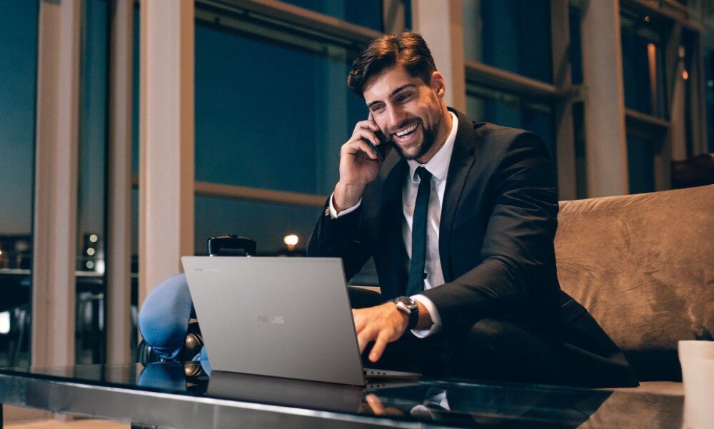 Laptop ASUS ExpertBook CX5403CMA-QM0137 Mężczyzna w garniturze siedzący na kanapie w przestrzeni biurowej; rozmawia przez telefon i pracuje na laptopie, łączność Wi-Fi 6E, porty