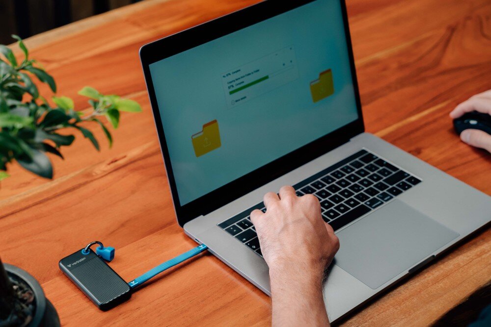 Dysk VERBATIM Pocket 1TB SSD Czarno-niebieski dysk leżący na biurku podłączony do laptopa, na którego ekranie pokazany jest proces przenoszenia plików interfejs prędkość podłączenie kabel kompatybilność