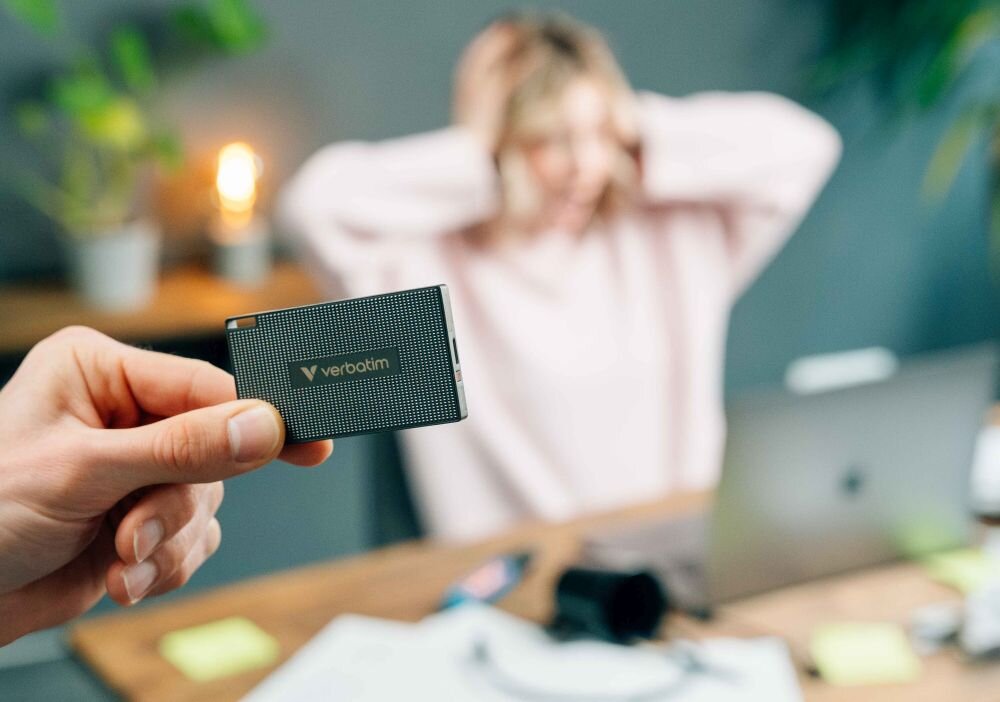 Dysk VERBATIM Metal Mini Zbliżenie dłoni trzymającej mały czarny dysk przenośny z widocznym napisem, w tle osoba siedząca przy biurku trzymająca ręce na głowie, opis