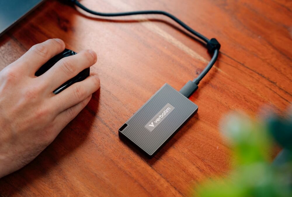 Dysk VERBATIM Metal Mini Dysk przenośny podłączony kablem do komputera leżący na drewnianym biurku obok dłoni korzystającej z myszy, interfejs, kabel