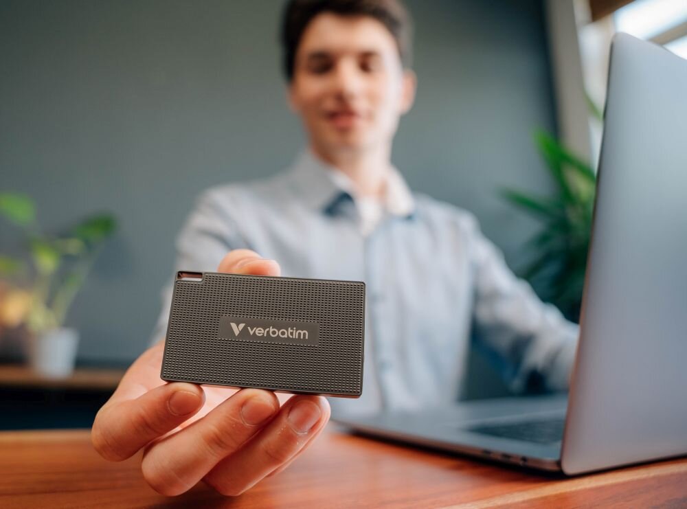 Dysk VERBATIM Metal Mini Mężczyzna siedzący przy biurku z laptopem trzymający w dłoni mały czarny dysk przenośny z widocznym napisem, pojemność, oprogramowanie, kompatybilność