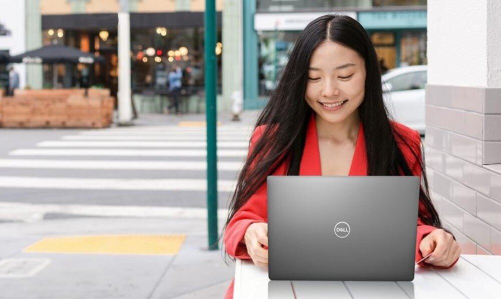 Laptop DELL Latitude 7340 Kobieta siedzi na zewnątrz przy stoliku z laptopem na kolanach, w tle przejście dla pieszych i miejskie kawiarnie, opis
