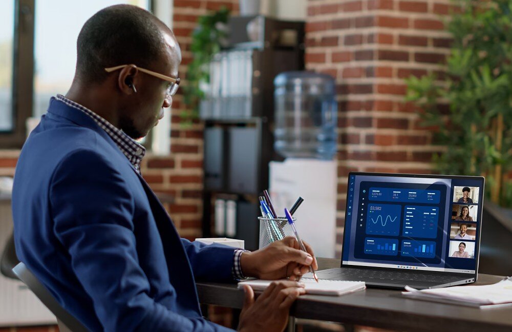 Laptop DELL Latitude 9450 Mężczyzna w okularach siedzi przy biurku w biurze typu open space, robi notatki i patrzy na laptopa, na którego ekranie widać wykresy i uczestników wideokonferencji, opis