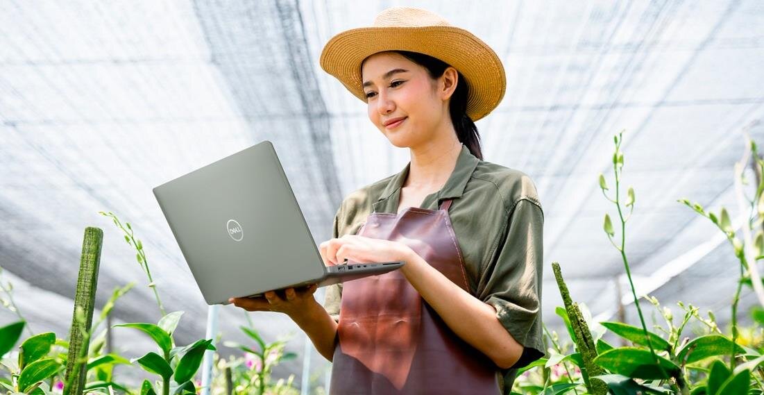 Kobieta w fartuchu trzyma laptop DELL Latitude 5440 w szklarni, otoczona roślinami. - Pamięć RAM LPDDR5X