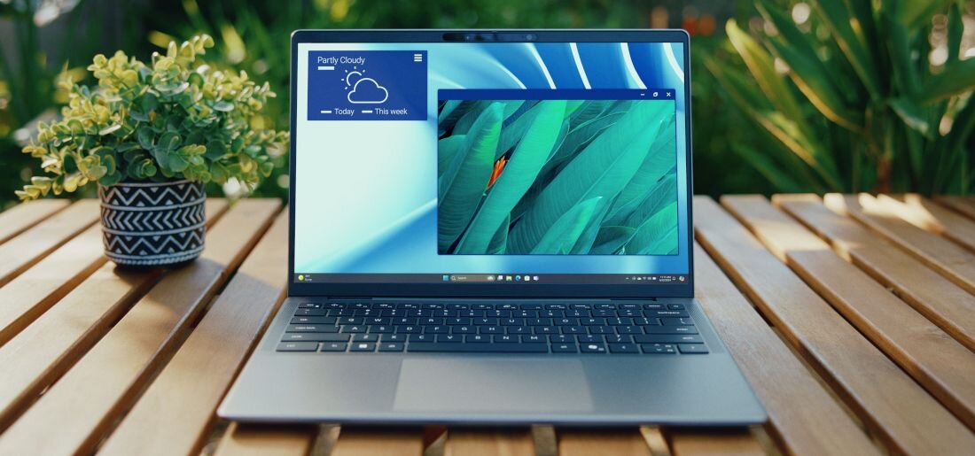 Laptop Dell Latitude z aplikacją pogodową Partly Cloudy wyświetlającą kolorową tapetę z liśćmi na drewnianym stole obok doniczki z rośliną.