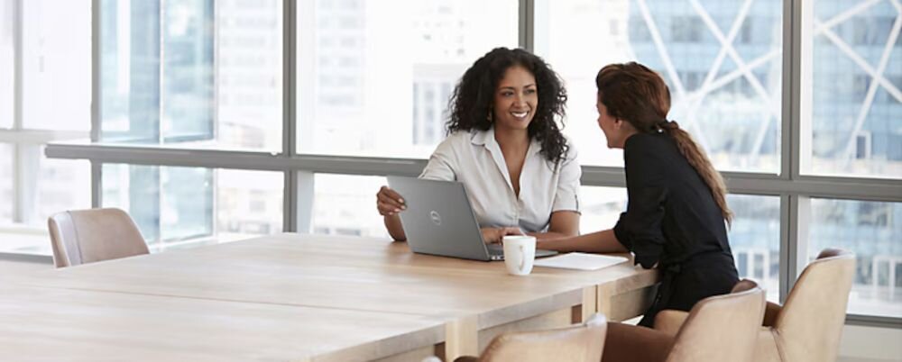 Dwie kobiety rozmawiające przy dużym stole konferencyjnym w nowoczesnym biurze z laptopem DELL Latitude na tle przeszklonych okien i miejskiego pejzażu. - Pamięć RAM DDR5