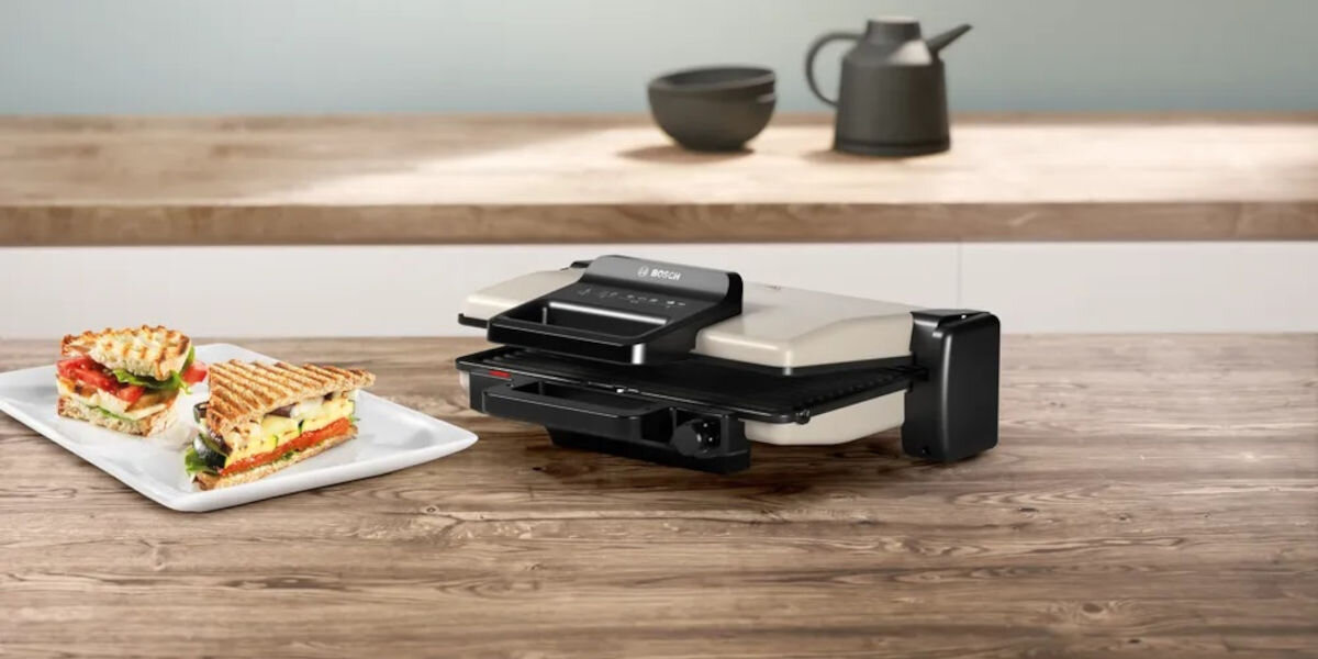 Grill elektryczny BOSCH TCG3302, kuchnia, pieczenie, smażenie, grillowanie, Grill BOSCH TCG3302 na blacie kuchennym, obok talerz z kanapkami panini, jasne tło kuchenne, drewno
