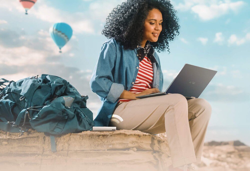 Router ASUS RT-AX57 Go AX3000 Uśmiechnięta kobieta siedząca na skale z laptopem na kolanach i białym routerem obok, na tle nieba z balonami na ogrzane powietrze