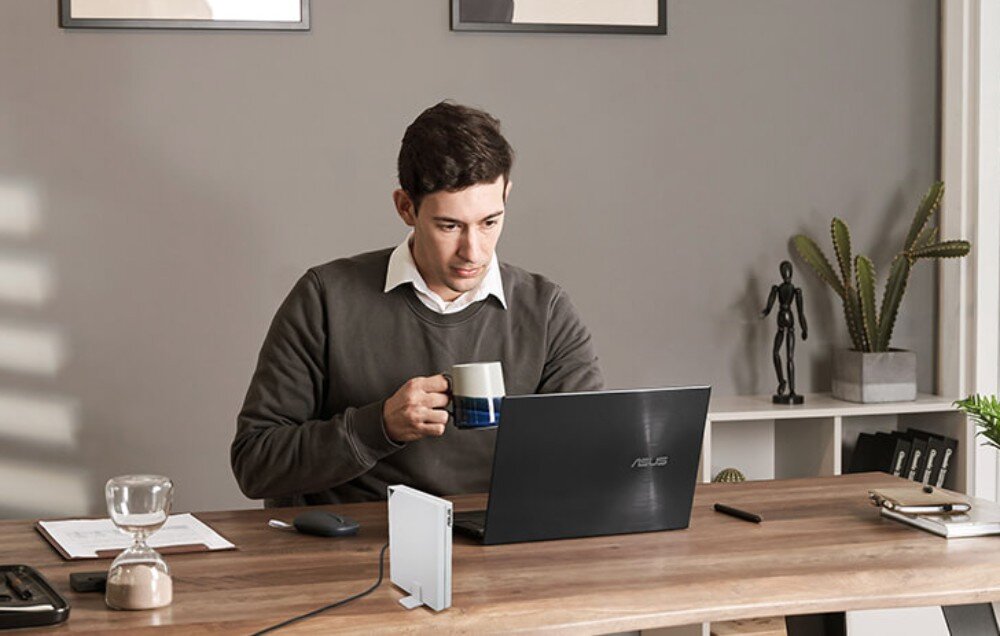 Router ASUS RT-AX57 Go AX3000 Mężczyzna pracujący przy biurku z laptopem i kubkiem kawy w dłoni, obok którego stoi biały router na rozkładanej nóżce