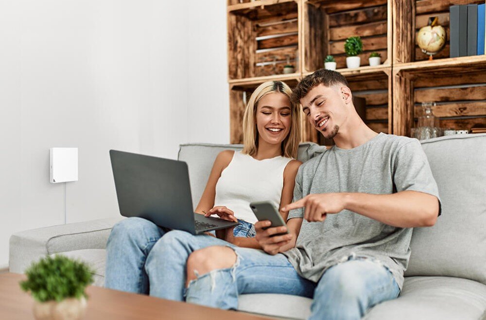 Router ASUS RT-AX57 Go AX3000 Młoda para siedząca na kanapie w salonie, korzystająca z laptopa i smartfona, z białym routerem zamontowanym na ścianie w tle