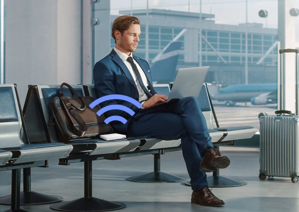 Router ASUS RT-AX57 Go AX3000 Mężczyzna w garniturze pracujący na laptopie w hali odlotów, z routerem leżącym na siedzeniu obok i grafiką symbolizującą sygnał Wi-Fi