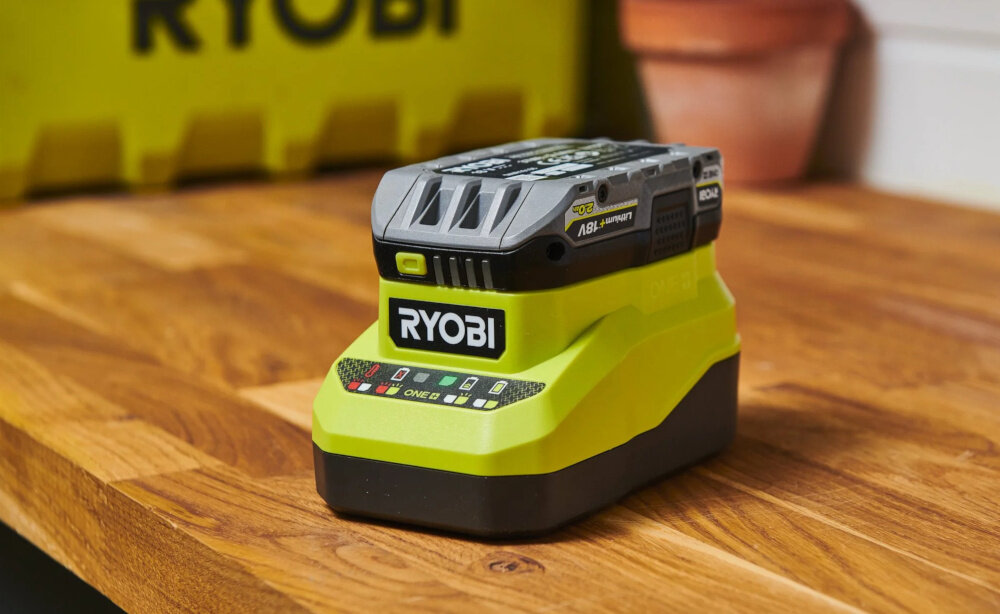 Na drewnianym blacie znajduje się akumulator z logo \'RYOBI\'. W tle widoczna jest żółta skrzynka z napisem \'RYOBI\'.