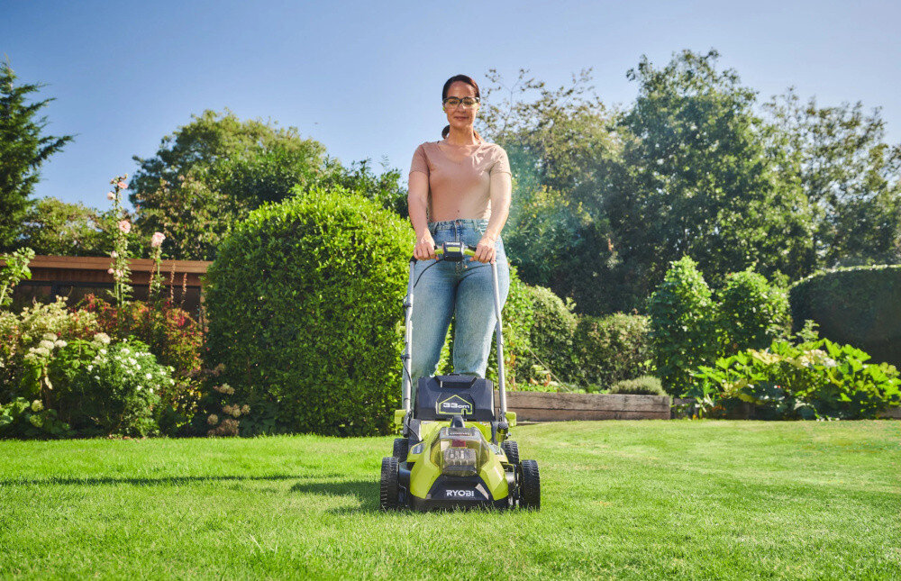 Osoba stoi na trawniku, prowadząc zieloną kosiarkę z widocznym logo \'RYOBI\'. W tle znajduje się ogród z krzewami i drzewami.