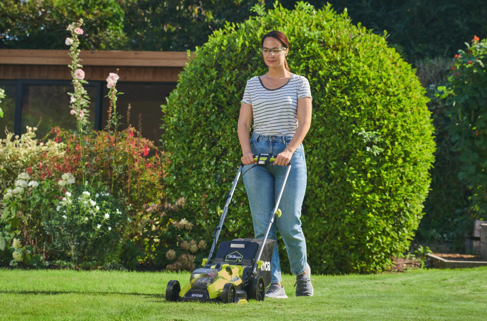 Osoba prowadzi kosiarkę akumulatorową marki RYOBI po trawniku w ogrodzie. W tle widoczne są krzewy i kwiaty.