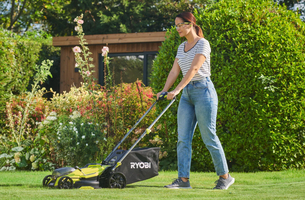 Osoba kosi trawę za pomocą kosiarki z widocznym logo \'RYOBI\'. W tle znajduje się zielony żywopłot i drewniana altana.