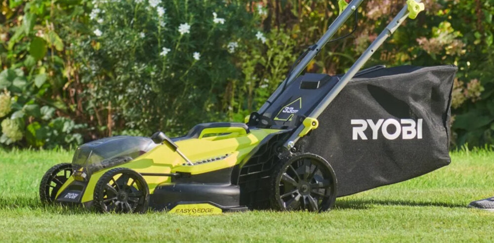 Na obrazie widoczna jest żółto-czarna kosiarka akumulatorowa z logo \'RYOBI\' na czarnym koszu na trawę. Kosiarka stoi na trawie, a w tle widać roślinność ogrodową.