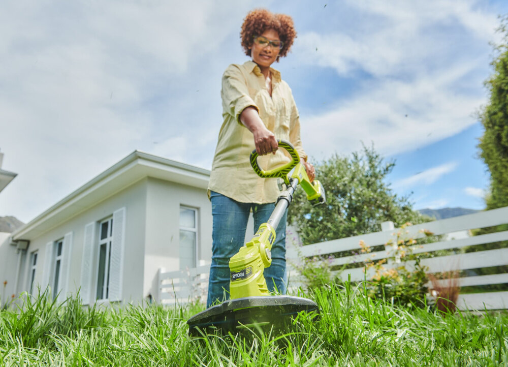 Osoba używa podkaszarki marki Ryobi na trawniku przed domem. W tle widoczny jest biały płot i budynek mieszkalny.