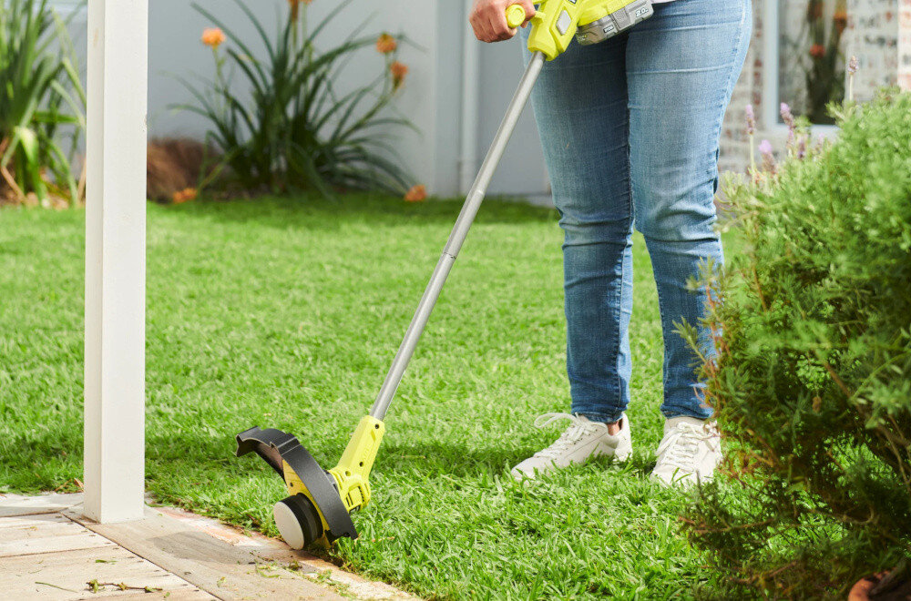 Osoba używa podkaszarki na trawniku w ogrodzie. W tle widoczne są rośliny i fragment budynku.