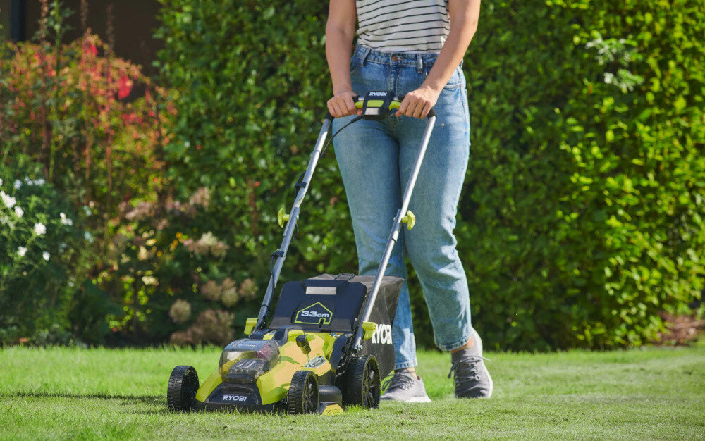 Osoba prowadzi kosiarkę akumulatorową marki RYOBI po trawniku. Na kosiarce widoczny jest napis \'33 cm\'.