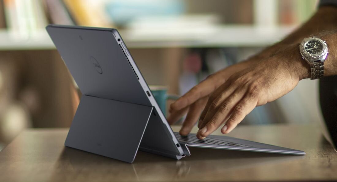 Zbliżenie dłoni mężczyzny z zegarkiem na nadgarstku dotykającej klawiatury tabletu DELL Latitude 7350 ze stojaczkiem w trybie laptopa, smukła konstrukcja w kolorze grafitowym, widoczne porty wentylacyjne, rozmazane tło z wnętrzem biurowym, urządzenie dla profesjonalnej pracy mobilnej​ - Opis serii Latitude 7350 