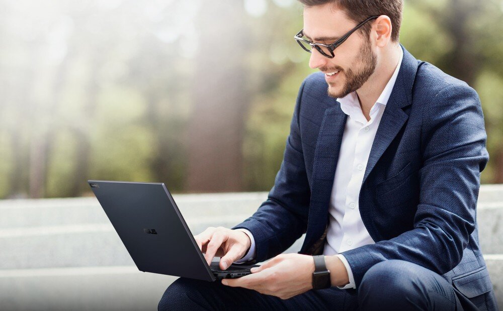Laptop ASUS ExpertBook B5404CMA-Q50339X Mężczyzna w garniturze i okularach przeciwsłonecznych siedzi na zewnątrz na schodach i korzysta z laptopa. Jest uśmiechnięty, trzyma dłonie na klawiaturze, a w tle widać rozmytą zieleń, przeznaczenie, procesor, wydajność, rdzenie, karta graficzna