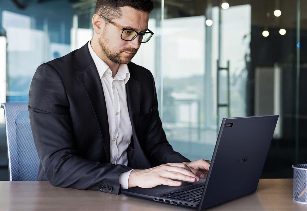Laptop ASUS ExpertBook B5404CMA-Q50339X Mężczyzna w eleganckim garniturze siedzi przy biurku w biurowcu i skupia się na pracy na laptopie. Szklane ściany i biurowe oświetlenie w tle, pamięć RAM, dysk, pojemność, interfejs