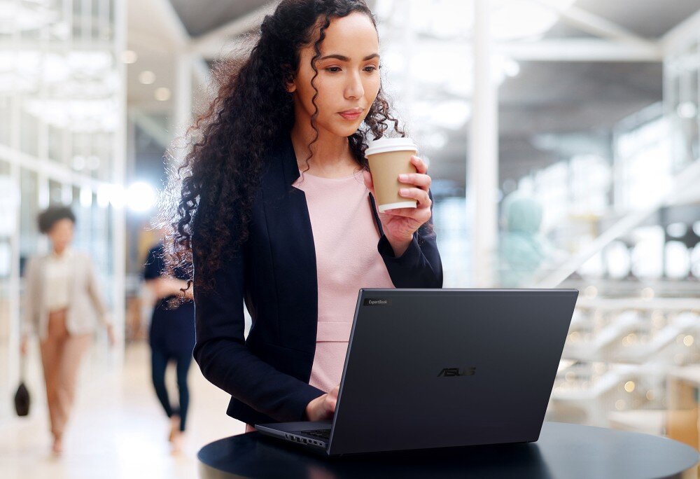 Laptop ASUS ExpertBook B5404CMA-Q50339X Kobieta w biznesowym stroju, z długimi, kręconymi włosami, stoi przy stoliku i korzysta z laptopa, trzymając jednocześnie kubek z kawą, obudowa, waga, wielkość, wytrzymałość