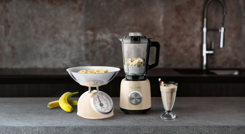 Blender kielichowy ETA Storio 901190010 Beżowy ciemne tło blender w kolorze beżowym stoi na ciemnym blacie srebrne pokrętło w kielichu zawartość obok w kieliszku koktajl obok blendera stoi beżowa waga stylowy design wysoka funkcjonalność eleganckie stalowe wykończenie solidny szklany kielich o pojemności 1,5 litra koktajle kremowe zupy sosy kruszenie lodu otwór w pokrywie dolewanie płynów podczas pracy precyzyjna kontrola 