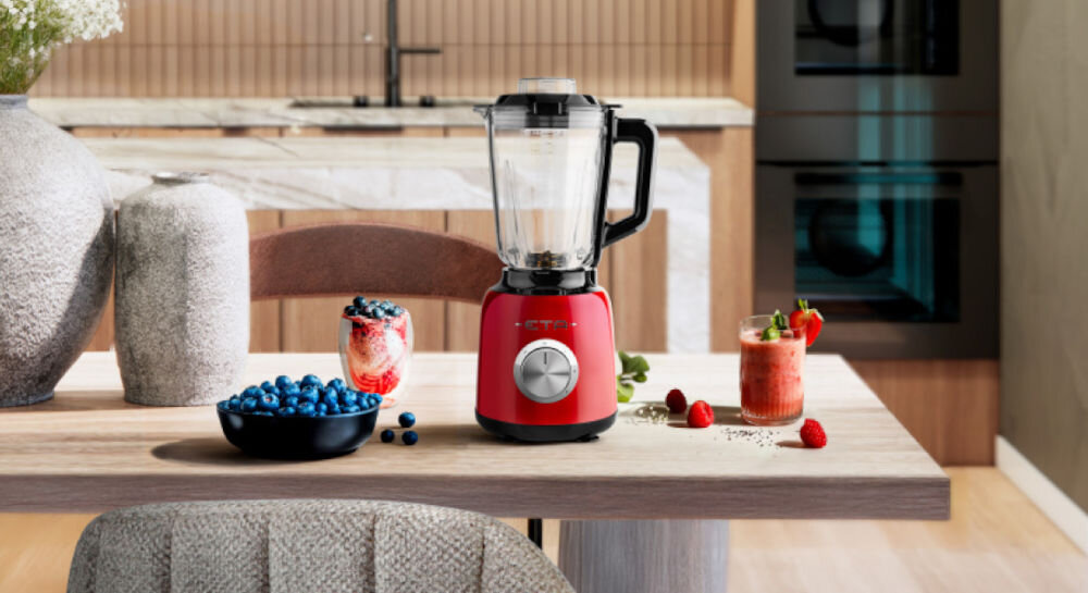 Blender kielichowy ETA Storio 901190020 Czerwony do koktajli czerwony blender stoi na blacie srebrne pokrętło czarna rączka kielich obok owoce w misce i napój w szklance 