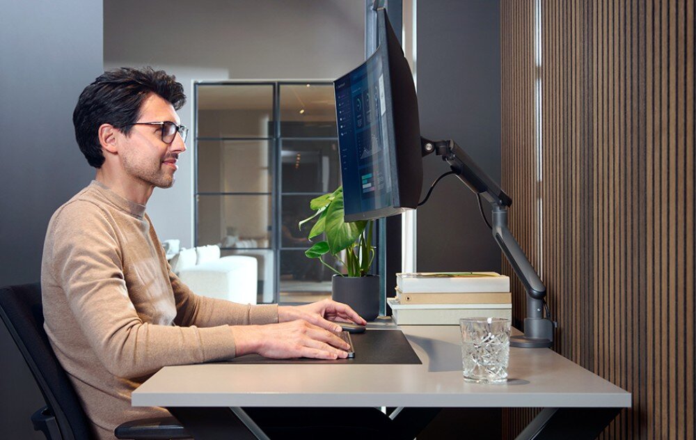 Uchwyt do monitora ONE FOR ALL DM7110 Mężczyzna w okularach siedzi przy biurku i patrzy na zakrzywiony monitor zamocowany na ruchomym ramieniu. Na blacie klawiatura, mysz, roślina w doniczce i szklanka wody