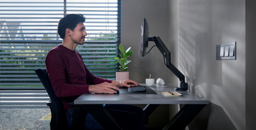 Uchwyt do monitora ONE FOR ALL DM4110 13-35 cali Czarny mężczyzna siedzący przy biurku z przymocowanym montorem na uchwycie ergonomia pracy, prawidłowa postawa, ramię monitora, redukcja zmęczenia