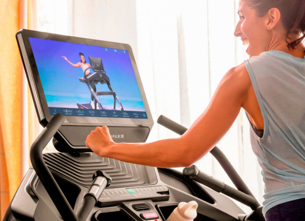 Bieżnia elektryczna BOWFLEX T56 2941W 20km-h osoba korzysta z wyświetlacza zbliżenie na wyświetlacz integracja z aplikacjami JRNY Explore the World Netflix Prime Video wzbogacenie treningów o multimedia oglądanie ulubionych filmów programów