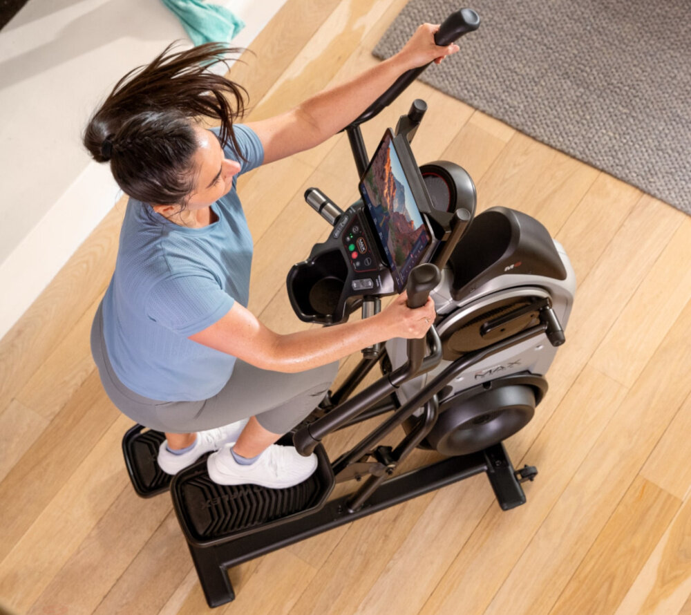 Orbitrek magnetyczno-powietrzny BOWFLEX M6i Max Trainer osoba ćwiczy na maszynie w pomieszczeniu widok z góry 16 poziomów oporu elektroniczna regulacja płynne zwiększanie lub zmniejszanie intensywności treningu dla osób początkujących i zaawansowanych