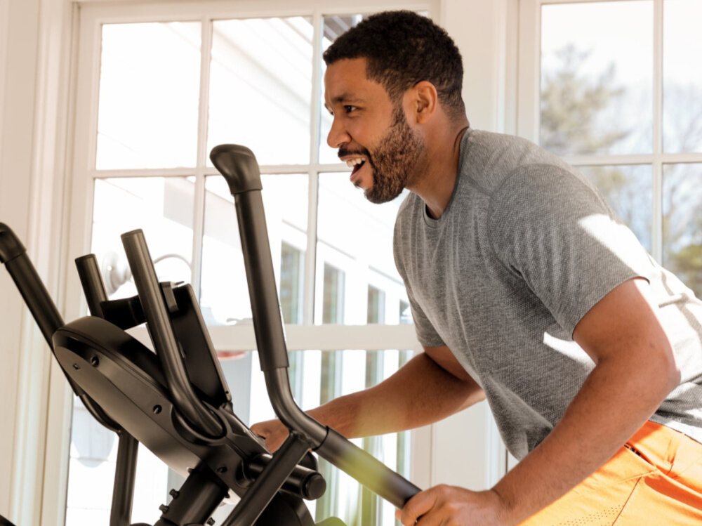 Orbitrek magnetyczno-powietrzny BOWFLEX M6i Max Trainer osoba ćwiczy na maszynie w pomieszczeniu zbliżenie na uchwyty monitorowanie tętna czujniki na uchwytach moduł Bluetooth 4.0 płynna transmisja danych