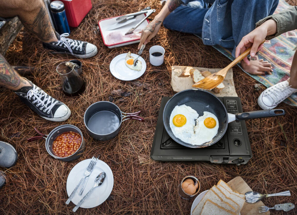 Kuchenka turystyczna EXTRALINK EX.39500 igliwie kilka osób siedzi wokół kuchenki na kuchence smażą się jajka wokół naczynia moc do 2500 W temperatura płomienia wynosi do 700°C krótszy czas oczekiwania większa efektywność gotowania