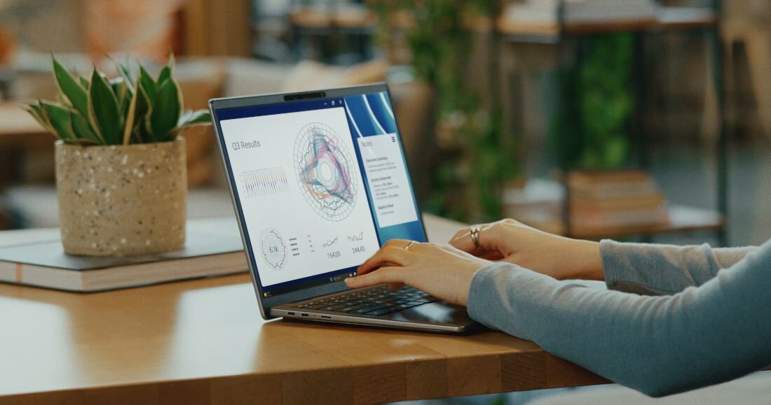 Dłonie pisząca na laptopie Dell Latitude z technologią ComfortView wyświetlającym aplikację 3D Results z kolorowymi wykresami kołowymi obok doniczki z rośliną. - Technologia ComfortView Plus z certyfikatem TÜV
