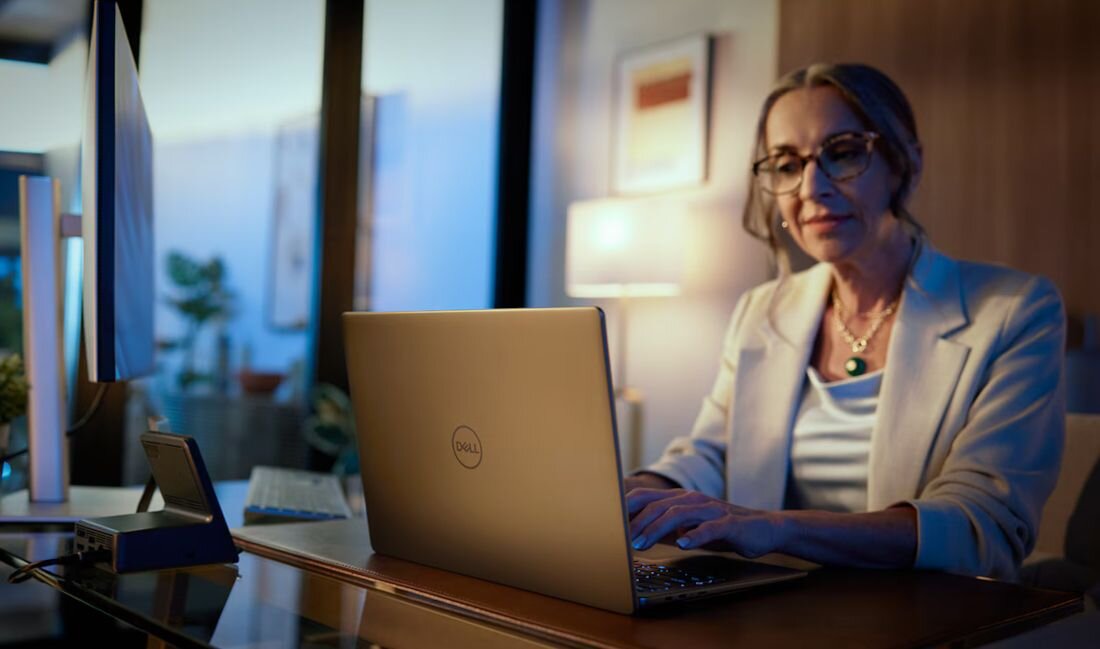 Kobieta w okularach i białym żakiecie używająca laptopa Dell Latitude w biurze z lampą i obrazami na ścianie w tle - Opis serii Latitude 7455