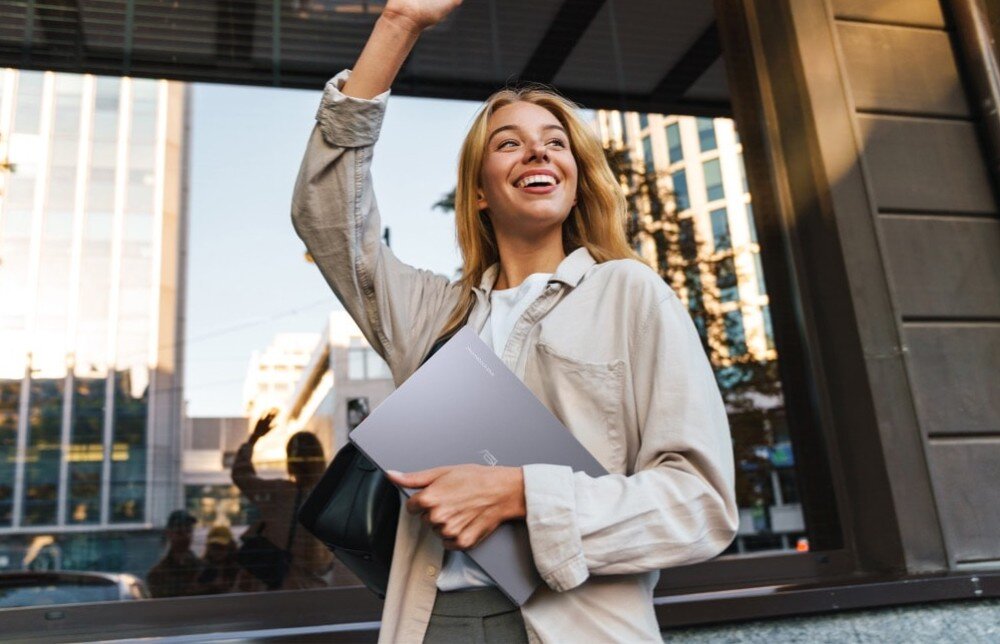 Laptop ASUS Chromebook Plus CB3402CVA Uśmiechnięta kobieta stojąca na tle budynków w centrum miasta. W jednej dłoni trzyma laptopa, drugą ręką macha do kogoś na powitanie, przeznaczenie, waga, wielkość, obudowa, wytrzymałość